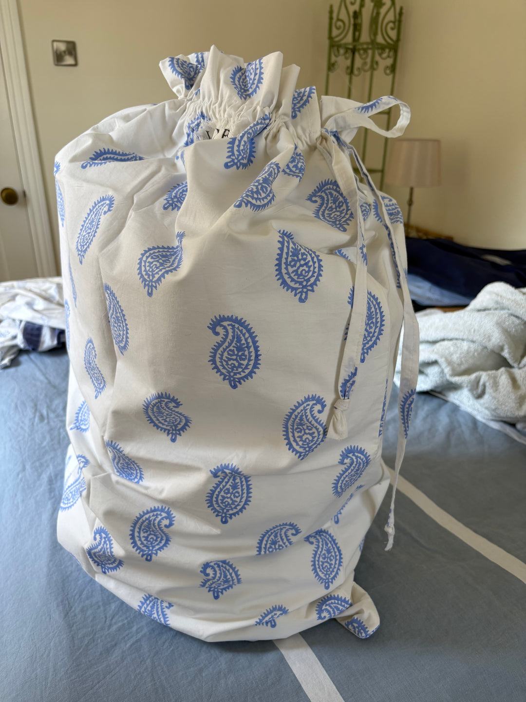 A white drawstring garment bag with a blue paisley pattern, photographed on a bed with a blue sheet and other clothes in the background.