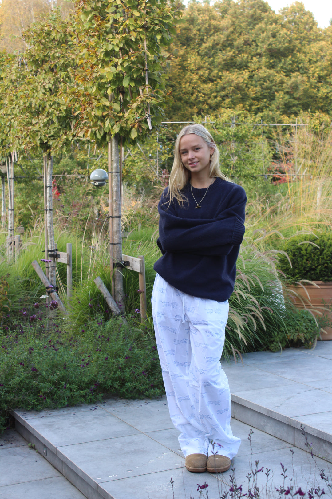 A woman wearing a dark navy blue sweatshirt and white wide-leg jeans stands on a paved path in a garden setting.