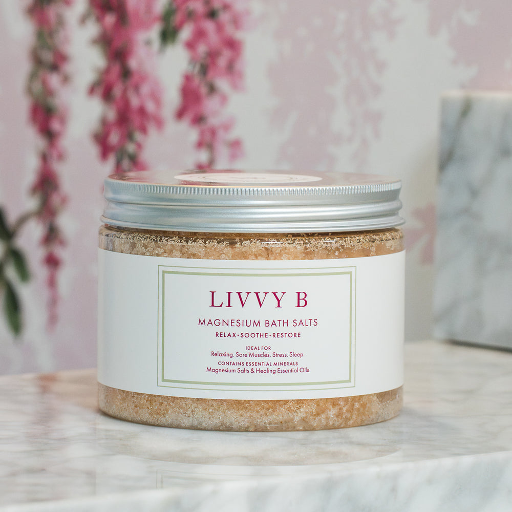 A jar of LIVVY B Magnesium Bath Salts with a white label and silver lid, set on a marble surface with a pink floral background.