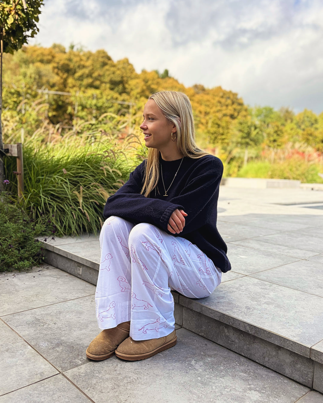 A young woman sits on stone steps outdoors, wearing a dark navy long-sleeve top, white lounge pants with a pink line-art pattern, and tan UGG slippers. The background features green plants and trees under a cloudy sky.