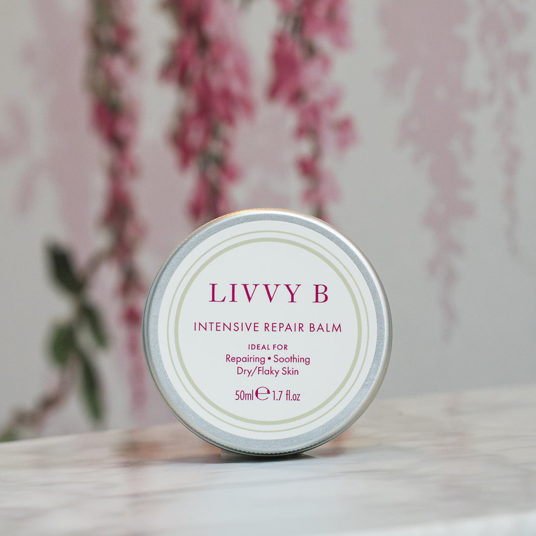 A round metal tin of LIVVY B Intensive Repair Balm sits on a light marble surface with a soft-focus floral wallpaper background.
