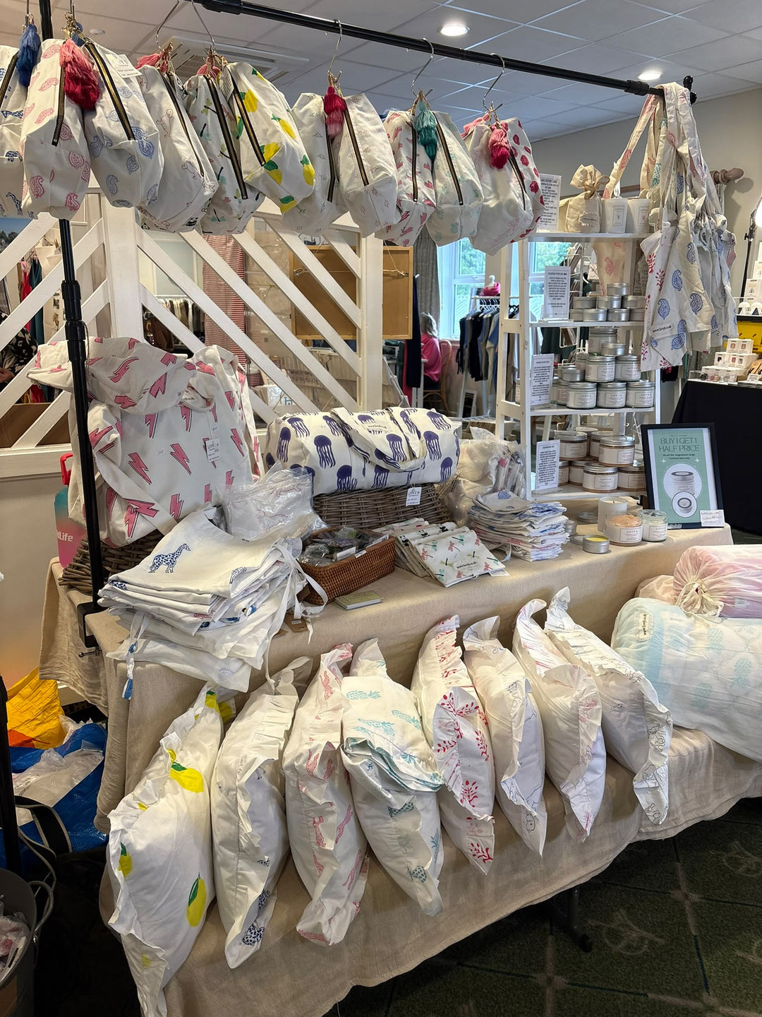 A retail display table filled with bedding and accessories, including patterned pillows, folded blankets, and hanging fabric bags, inside a boutique store.