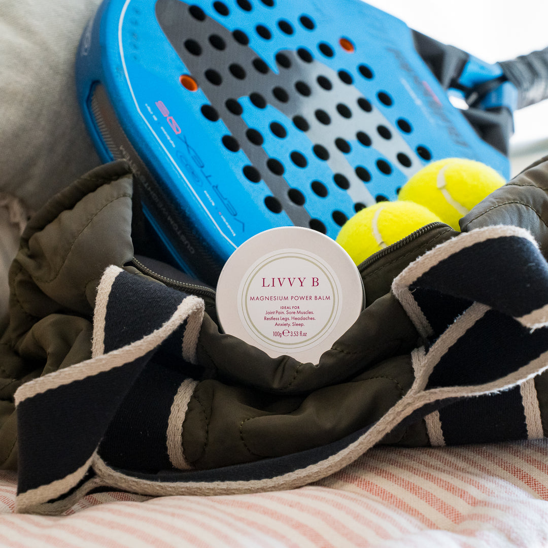 A round white tin of LIVVY B Magnesium Power Balm sits in front of a dark navy and cream striped quilted bag, with a blue tennis paddle and yellow tennis balls in the background.