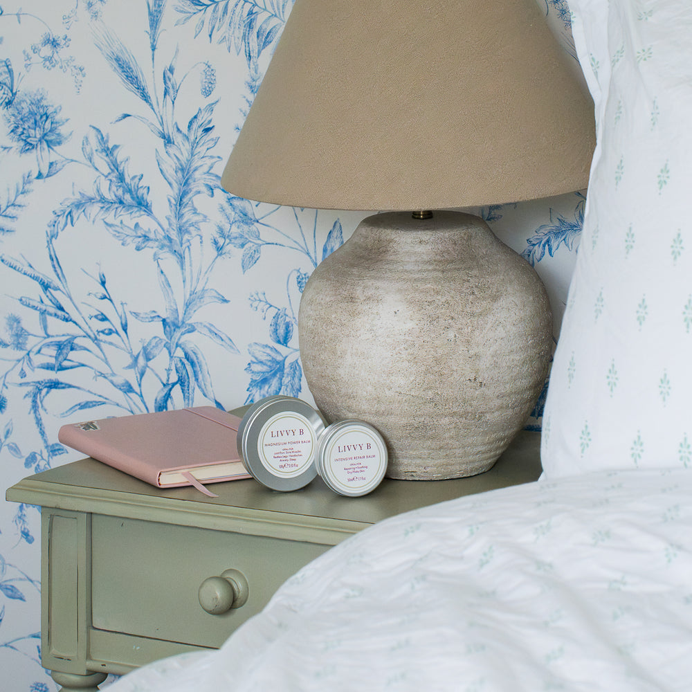 Two small round tin candles with silver lids and white labels reading 'LIVVY B ' are styled on a pale sage green bedside table next to a pink notebook and a textured beige table lamp, set against blue and white floral wallpaper.