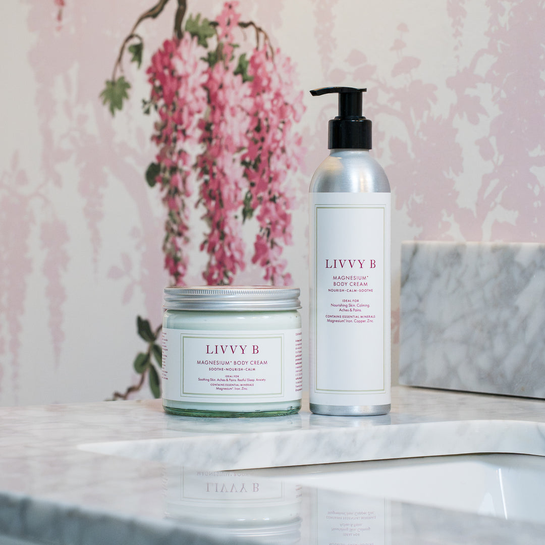 Two Livvy B skincare products, a magnesium body cream jar and a magnesium body lotion bottle, displayed on a white marble surface against a pink floral wallpaper background.