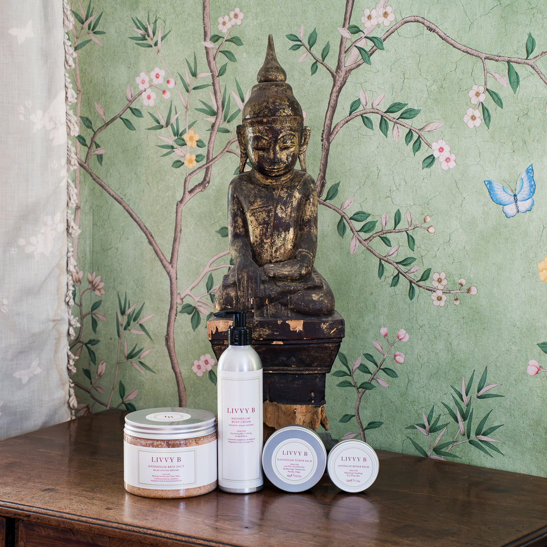 Four LIVVY B  products arranged on a wooden table in front of a decorative Buddha statue and green floral wallpaper. The set includes a round tin candle, a tall white bottle with a black pump, and two small round jars with white lids.