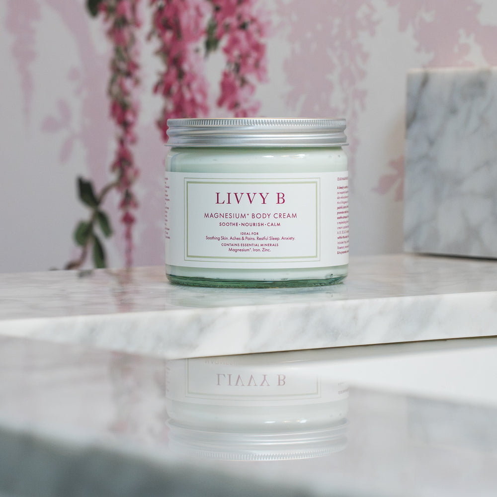 A jar of Livvy B Magnesium Body Cream sits on a white marble surface with a pink floral wallpaper background.