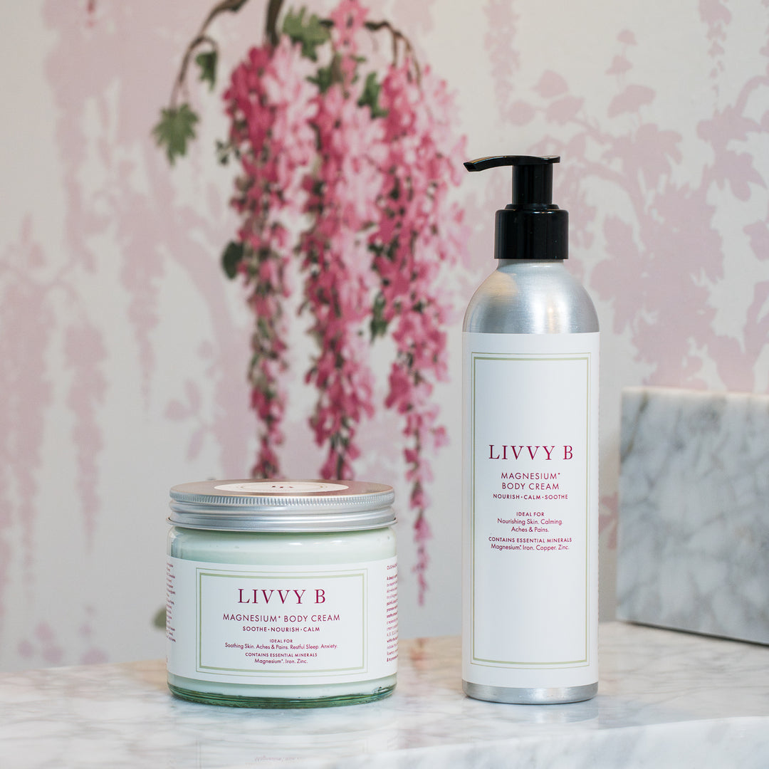 Two LIVVY B body care products on a marble surface: a small glass jar of Nourishing Body Cream with a silver lid and a tall silver pump bottle of Magnesium Body Cream, set against a pink floral wallpaper background.
